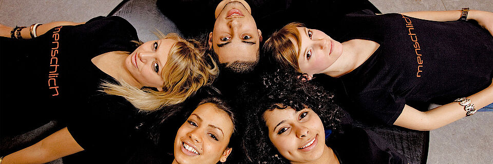 Fünf Jugendliche liegen Kopf an Kopf im Kreis. Sie lachen und wirken vergnügt und herzlich. Alle tragen ein schwarzes T-Shirt mit der Aufschrift 'menschlich'. Für das Jugendrotkreuz